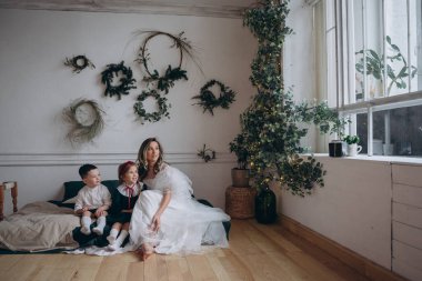 Küçük kızı ve oğluyla birlikte Noel süslemeleri olan bir odada beyaz bir bohem elbisesi giymiş güzel bir kadın.