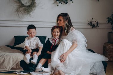 Küçük kızı ve oğluyla birlikte Noel süslemeleri olan bir odada beyaz bir bohem elbisesi giymiş güzel bir kadın.