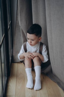 Perdelerle pencerenin yanında oturan neşeli elbiseli yakışıklı çocuk.