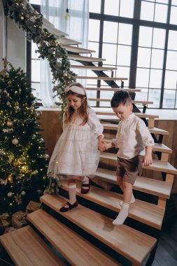 Erkek kardeşiyle bohem beyaz elbiseler giymiş güzel bir kız pencerenin arkasındaki ahşap merdivenlerden iniyor.