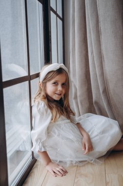 Bohem beyaz elbiseli güzel bir kız pencerenin yanında oturmuş kameraya poz veriyor.