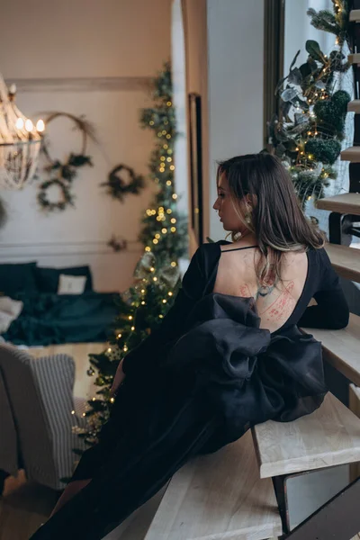 beautiful woman with tattoos in the dark fancy dress sitting on stairs and posing for camera in a room with christmas decorations