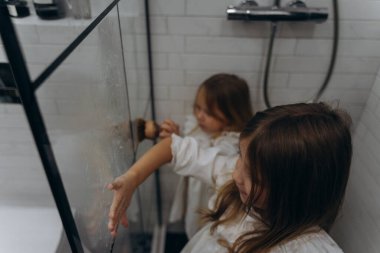 Beyaz pamuklu gecelikler giymiş şirin küçük kızlar ellerini sabunla yıkıyor ve hafif bir banyoda duşun camını köpükle kaplıyorlar.