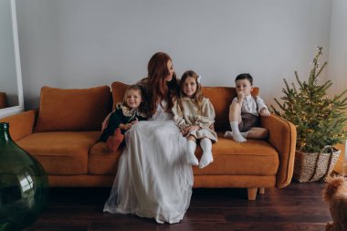 Beyaz bohem bir elbise giymiş güzel kızıl saçlı kadın küçük kızları ve oğluyla birlikte Noel arifesinde kanepede oturuyor. 