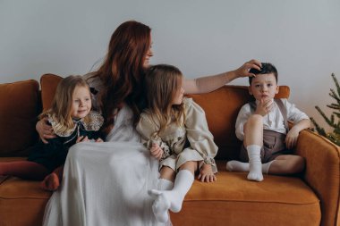 Beyaz bohem bir elbise giymiş güzel kızıl saçlı kadın küçük kızları ve oğluyla birlikte Noel arifesinde kanepede oturuyor. 