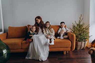 Beyaz bohem bir elbise giymiş güzel kızıl saçlı kadın küçük kızları ve oğluyla birlikte Noel arifesinde kanepede oturuyor. 