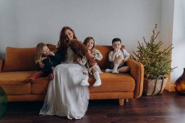 Beyaz bohem bir elbise giymiş güzel kızıl saçlı kadın küçük kızları ve oğluyla birlikte Noel arifesinde kanepede oturuyor. 