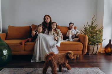 Beyaz bohem bir elbise giymiş güzel kızıl saçlı kadın küçük kızları ve oğluyla birlikte Noel arifesinde kanepede oturuyor. 