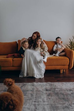 Beyaz bohem bir elbise giymiş güzel kızıl saçlı kadın küçük kızları ve oğluyla birlikte Noel arifesinde kanepede oturuyor. 
