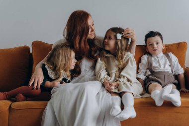 Beyaz bohem bir elbise giymiş güzel kızıl saçlı kadın küçük kızları ve oğluyla birlikte Noel arifesinde kanepede oturuyor. 