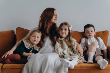 Beyaz bohem bir elbise giymiş güzel kızıl saçlı kadın küçük kızları ve oğluyla birlikte Noel arifesinde kanepede oturuyor. 