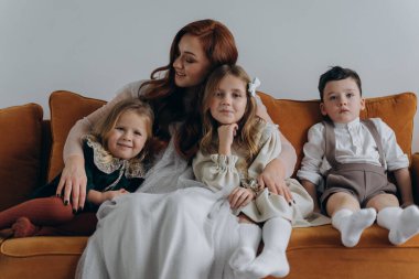 Beyaz bohem bir elbise giymiş güzel kızıl saçlı kadın küçük kızları ve oğluyla birlikte Noel arifesinde kanepede oturuyor. 