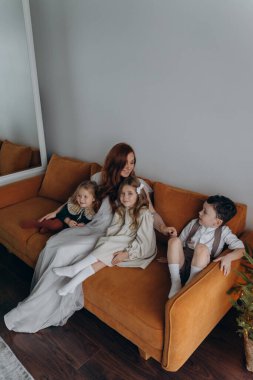 Beyaz bohem bir elbise giymiş güzel kızıl saçlı kadın küçük kızları ve oğluyla birlikte Noel arifesinde kanepede oturuyor. 