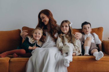 Beyaz bohem bir elbise giymiş güzel kızıl saçlı kadın küçük kızları ve oğluyla birlikte Noel arifesinde kanepede oturuyor. 