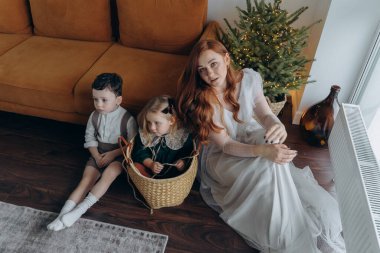 Küçük kızı ve oğluyla birlikte beyaz bohem bir elbise giymiş güzel bir kızıl kadın Noel arifesinde birlikte vakit geçiriyorlar ve kız sepette oturuyor. 