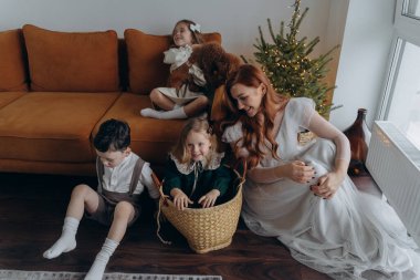 Kızları ve oğluyla birlikte beyaz bohem elbiseler giymiş güzel bir kızıl kadın Noel arifesinde birlikte vakit geçiriyorlar ve en küçük kız sepette oturuyor.