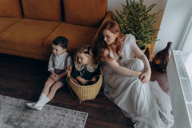 Küçük kızı ve oğluyla birlikte beyaz bohem bir elbise giymiş güzel bir kızıl kadın Noel arifesinde birlikte vakit geçiriyorlar ve kız sepette oturuyor. 