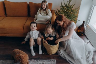 Kızları ve oğluyla birlikte beyaz bohem elbiseler giymiş güzel bir kızıl kadın Noel arifesinde birlikte vakit geçiriyorlar ve en küçük kız sepette oturuyor.