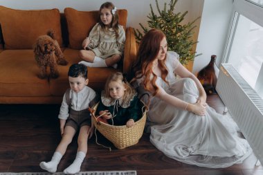 Kızları ve oğluyla birlikte beyaz bohem elbiseler giymiş güzel bir kızıl kadın Noel arifesinde birlikte vakit geçiriyorlar ve en küçük kız sepette oturuyor.