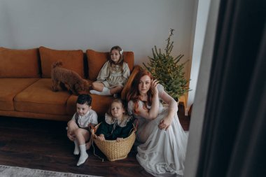 Kızları ve oğluyla birlikte beyaz bohem elbiseler giymiş güzel bir kızıl kadın Noel arifesinde birlikte vakit geçiriyorlar ve en küçük kız sepette oturuyor.