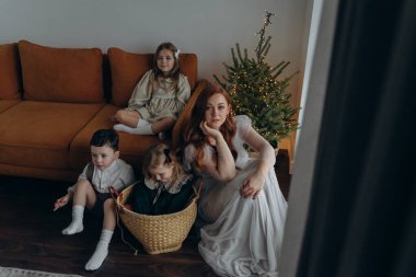Kızları ve oğluyla birlikte beyaz bohem elbiseler giymiş güzel bir kızıl kadın Noel arifesinde birlikte vakit geçiriyorlar ve en küçük kız sepette oturuyor.