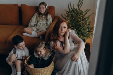 Kızları ve oğluyla birlikte beyaz bohem elbiseler giymiş güzel bir kızıl kadın Noel arifesinde birlikte vakit geçiriyorlar ve en küçük kız sepette oturuyor.