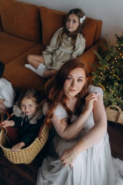 Kızları ve oğluyla birlikte beyaz bohem elbiseler giymiş güzel bir kızıl kadın Noel arifesinde birlikte vakit geçiriyorlar ve en küçük kız sepette oturuyor.