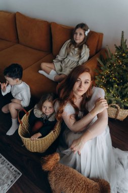 Kızları ve oğluyla birlikte beyaz bohem elbiseler giymiş güzel bir kızıl kadın Noel arifesinde birlikte vakit geçiriyorlar ve en küçük kız sepette oturuyor.