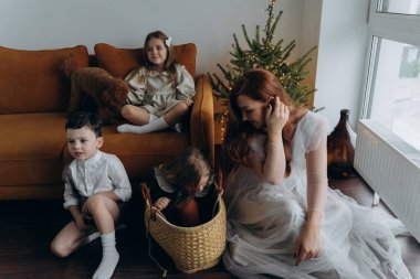 Kızları ve oğluyla birlikte beyaz bohem elbiseler giymiş güzel bir kızıl kadın Noel arifesinde birlikte vakit geçiriyorlar ve en küçük kız sepette oturuyor.