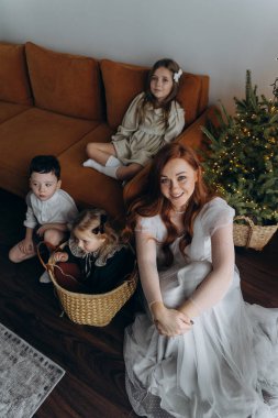 Kızları ve oğluyla birlikte beyaz bohem elbiseler giymiş güzel bir kızıl kadın Noel arifesinde birlikte vakit geçiriyorlar ve en küçük kız sepette oturuyor.