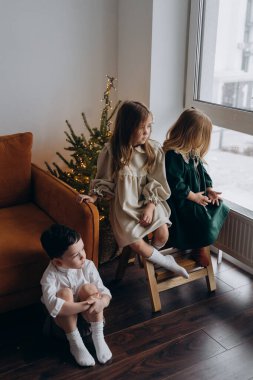 Noel arifesinde erkek kardeşleriyle birlikte pencerenin kenarında oturan bohem elbiseli güzel küçük kızlar.