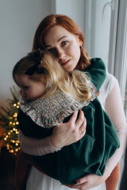 Beyaz bohem bir elbise giymiş güzel kızıl saçlı kadın ve küçük kızı pencerenin yanında birbirlerine sarılıyorlar.