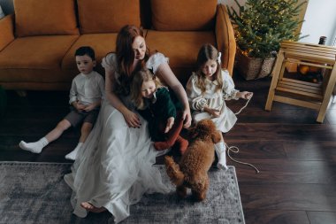 Oğlu ve küçük tatlı kızlarıyla birlikte beyaz bohem elbiseler giymiş güzel kızıl saçlı kadın kanepedeki evlerinde köpekleriyle vakit geçiriyorlar.