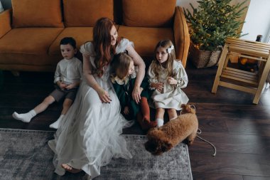 Oğlu ve küçük tatlı kızlarıyla birlikte beyaz bohem elbiseler giymiş güzel kızıl saçlı kadın kanepedeki evlerinde köpekleriyle vakit geçiriyorlar.