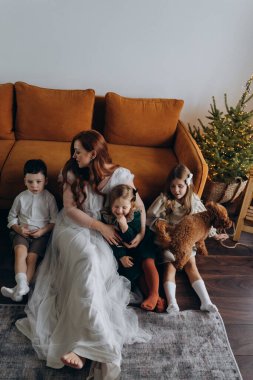 Oğlu ve küçük tatlı kızlarıyla birlikte beyaz bohem elbiseler giymiş güzel kızıl saçlı kadın kanepedeki evlerinde köpekleriyle vakit geçiriyorlar.