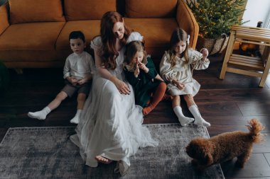 Oğlu ve küçük tatlı kızlarıyla birlikte beyaz bohem elbiseler giymiş güzel kızıl saçlı kadın kanepedeki evlerinde köpekleriyle vakit geçiriyorlar.