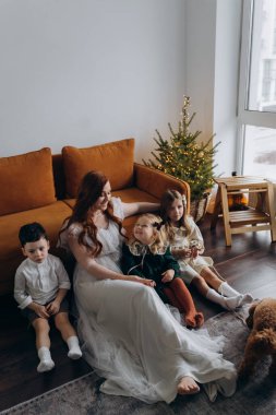 Oğlu ve küçük tatlı kızlarıyla birlikte beyaz bohem elbiseler giymiş güzel kızıl saçlı kadın kanepedeki evlerinde köpekleriyle vakit geçiriyorlar.