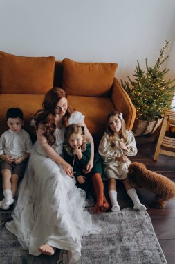 Oğlu ve küçük tatlı kızlarıyla birlikte beyaz bohem elbiseler giymiş güzel kızıl saçlı kadın kanepedeki evlerinde köpekleriyle vakit geçiriyorlar.