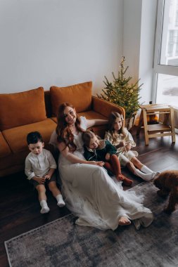 Oğlu ve küçük tatlı kızlarıyla birlikte beyaz bohem elbiseler giymiş güzel kızıl saçlı kadın kanepedeki evlerinde köpekleriyle vakit geçiriyorlar.