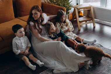 Oğlu ve küçük tatlı kızlarıyla birlikte beyaz bohem elbiseler giymiş güzel kızıl saçlı kadın kanepedeki evlerinde köpekleriyle vakit geçiriyorlar.