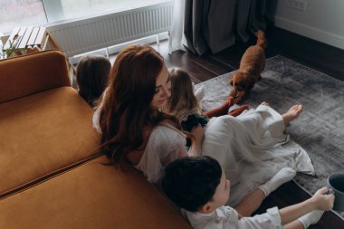 Oğlu ve küçük tatlı kızlarıyla birlikte beyaz bohem elbiseler giymiş güzel kızıl saçlı kadın kanepedeki evlerinde köpekleriyle vakit geçiriyorlar.