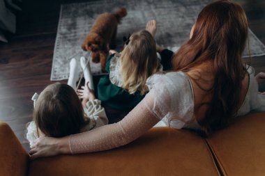 Oğlu ve küçük tatlı kızlarıyla birlikte beyaz bohem elbiseler giymiş güzel kızıl saçlı kadın kanepedeki evlerinde köpekleriyle vakit geçiriyorlar.