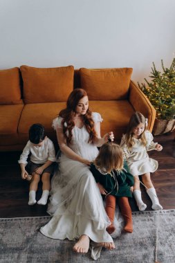 Oğlu ve tatlı küçük kızlarıyla birlikte beyaz bohem elbiseler giymiş güzel kızıl saçlı kadın kanepede birlikte vakit geçiriyorlar.