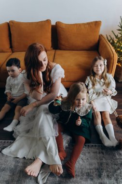 Oğlu ve küçük tatlı kızlarıyla birlikte beyaz bohem elbiseler giymiş güzel kızıl saçlı kadın kanepedeki evlerinde köpekleriyle vakit geçiriyorlar.