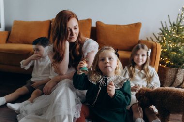 Oğlu ve küçük tatlı kızlarıyla birlikte beyaz bohem elbiseler giymiş güzel kızıl saçlı kadın kanepedeki evlerinde köpekleriyle vakit geçiriyorlar.