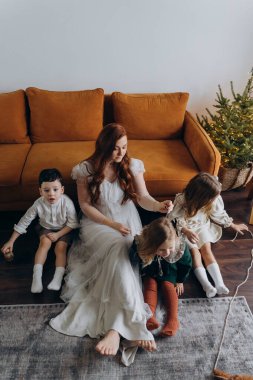 Oğlu ve küçük tatlı kızlarıyla birlikte beyaz bohem elbiseler giymiş güzel kızıl saçlı kadın kanepedeki evlerinde köpekleriyle vakit geçiriyorlar.