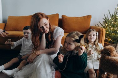 Oğlu ve küçük tatlı kızlarıyla birlikte beyaz bohem elbiseler giymiş güzel kızıl saçlı kadın kanepedeki evlerinde köpekleriyle vakit geçiriyorlar.