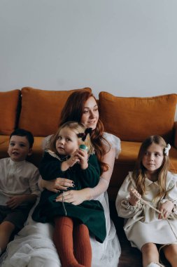 Oğlu ve tatlı küçük kızlarıyla birlikte beyaz bohem elbiseler giymiş güzel kızıl saçlı kadın kanepede birlikte vakit geçiriyorlar.