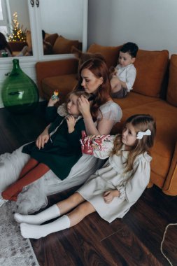 Oğlu ve tatlı küçük kızlarıyla birlikte beyaz bohem elbiseler giymiş güzel kızıl saçlı kadın kanepede birlikte vakit geçiriyorlar.