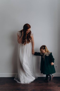 Beyaz bohem bir elbise giymiş güzel kızıl saçlı kadın küçük kızının elini tutuyor ve kameraya birlikte poz veriyor. 